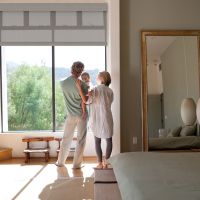 Couple holding a child in a bedroom with motorized shades partially lowered over large windows offering outdoor views.